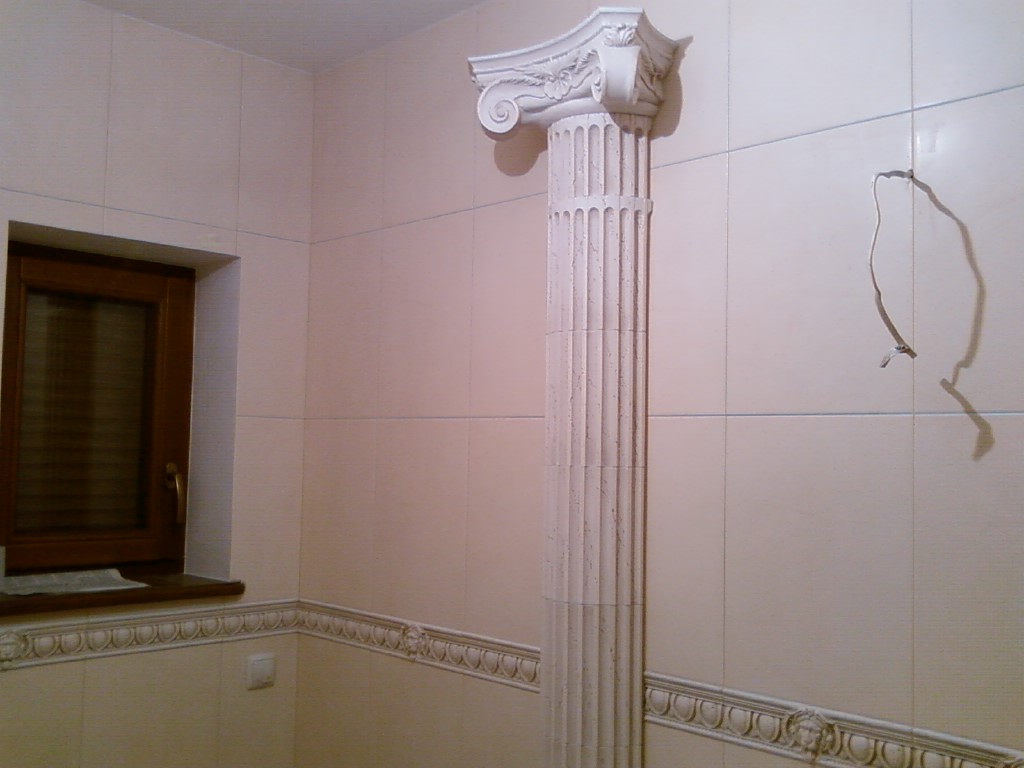 Wykończona łazienka z beżowymi płytkami, dekoracyjną kolumną w stylu greckim i drewnianym oknem. Widoczny wystający kabel ze ściany.